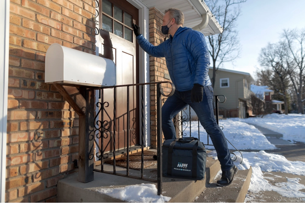 White Male Volunteer Blue Jacket Knocking on Door with Meal Delivery